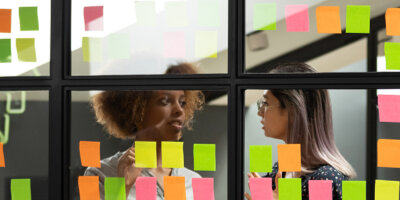 Header Image: Two female colleagues discuss their scrum board that is covered in post it notes