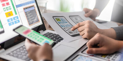 Two people lean over a laptop comparing a drawn mock up of a mobile app to the digital mock up, to the actual mobile experience on a cellphone