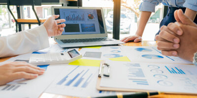 A table is strewn with reports showing pie charts and other data. A hand points at a laptop where more reporting is displayed.