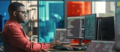 A man sits at his computer typing. Code is displayed across his three monitors.