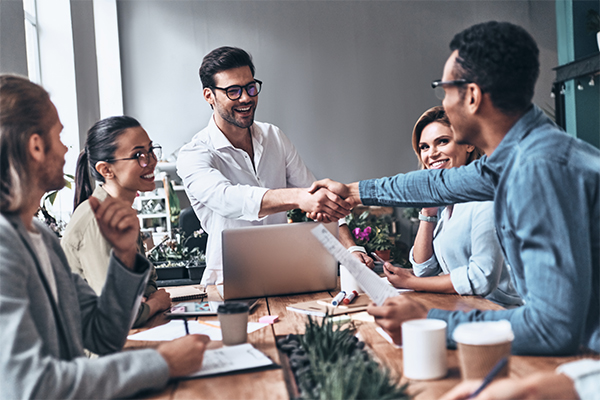 A diverse group of account executives sit around a table. One leans across to shake hands with a new client.