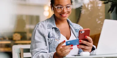 A woman who has been browsing on her laptop, holds a credit card up and completes an order on her phone.