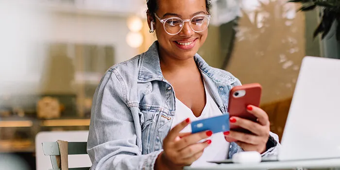 A woman who has been browsing on her laptop, holds a credit card up and completes an order on her phone.