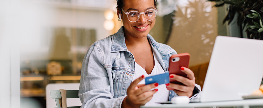 A woman who has been browsing on her laptop, holds a credit card up and completes an order on her phone.