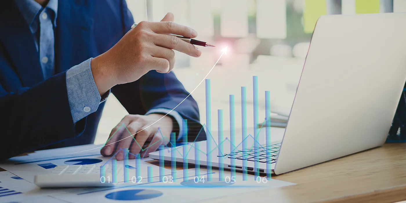 A man sitting at his desk with his laptop open draws a curve above a hologram of a bar graph, showing how it has increased over time.